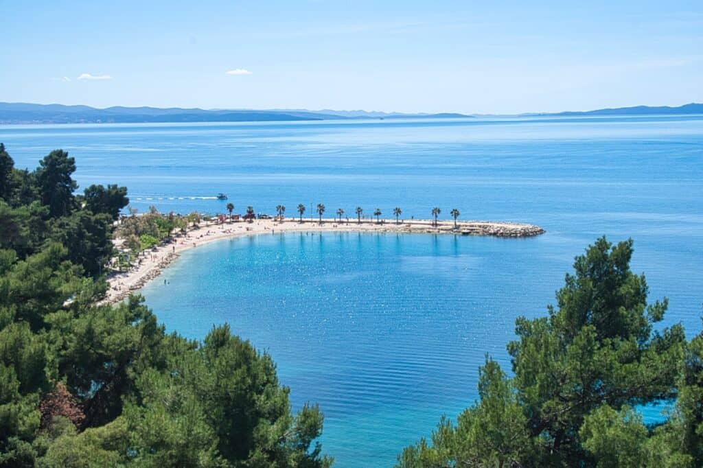 Der schöne Kasjuni Strand in Split aus der Vogelperspektive. Man sieht die Bucht und die Palmen sowie ein Boot dahinter fahren