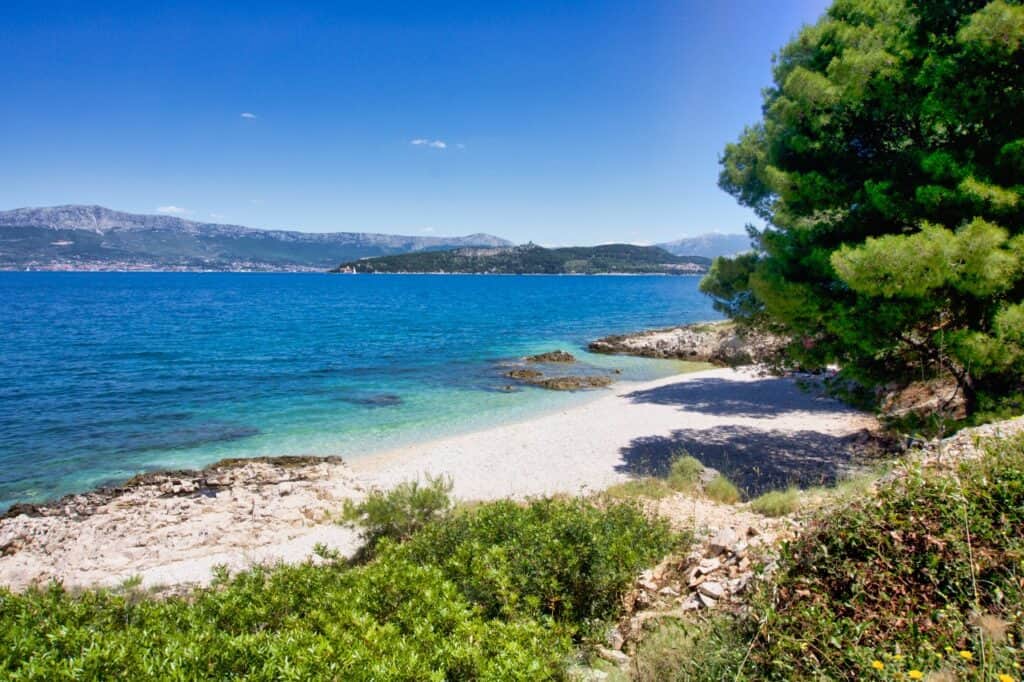 Der Glorija Beach mit einigen Bäumen und hellblauem Wasser auf der Insel Čiovo in der Nähe von Trogir